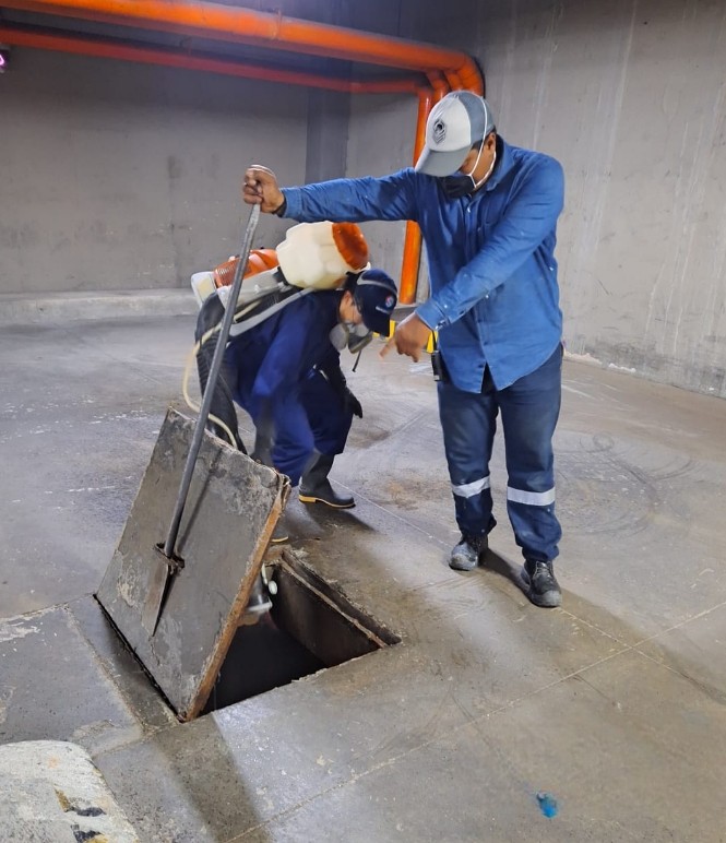 Dos trabajadores de Sergecol Saneamiento Ambiental abren una tapa de acceso a un conducto en un parqueadero subterráneo; uno sostiene la tapa con una barra mientras el otro revisa el interior del conducto. Ambos usan uniforme, casco y mascarilla de protección.